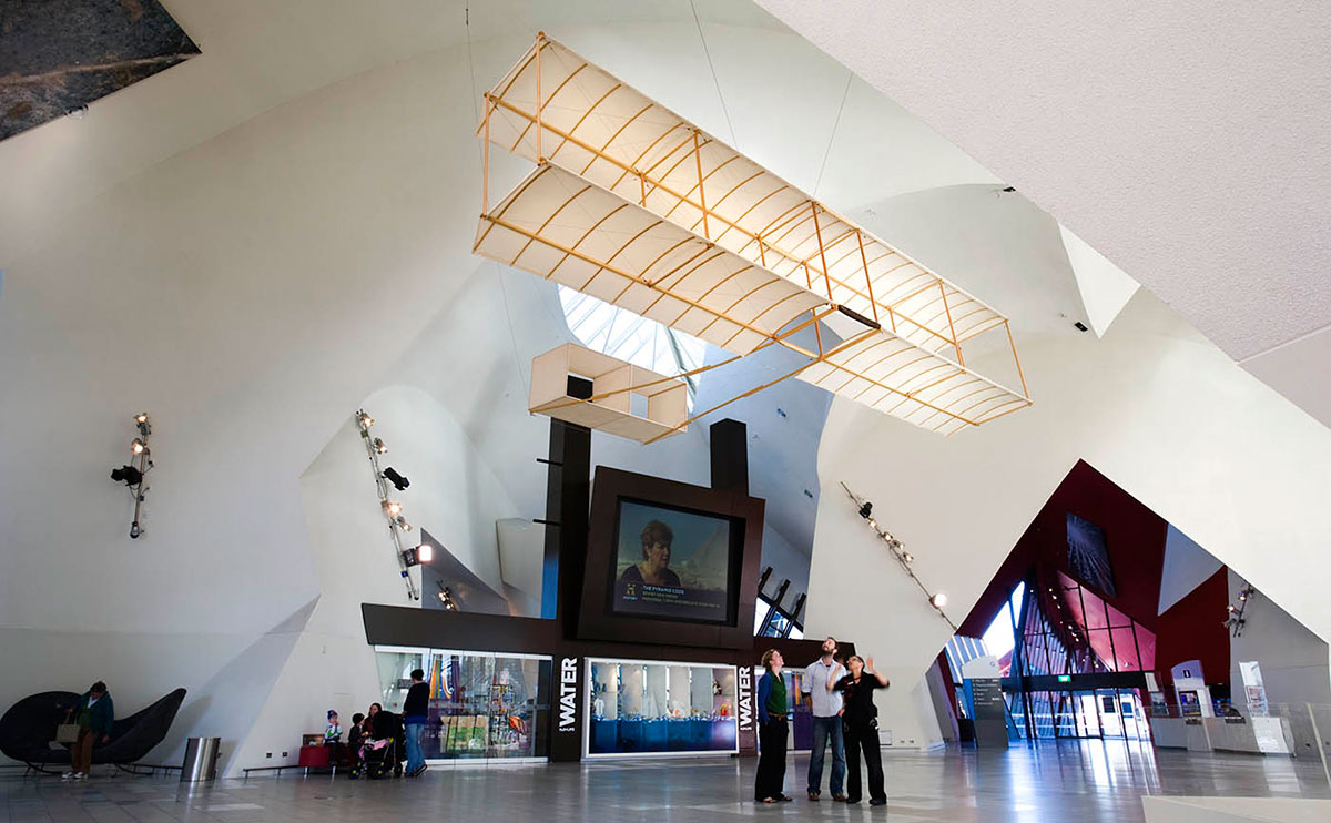 A replica of a biplane (two wing) glider suspended from the ceiling of a hall in a museum. The glider is made from wood and fabric. A small group of people stand under the glider looking up at at.