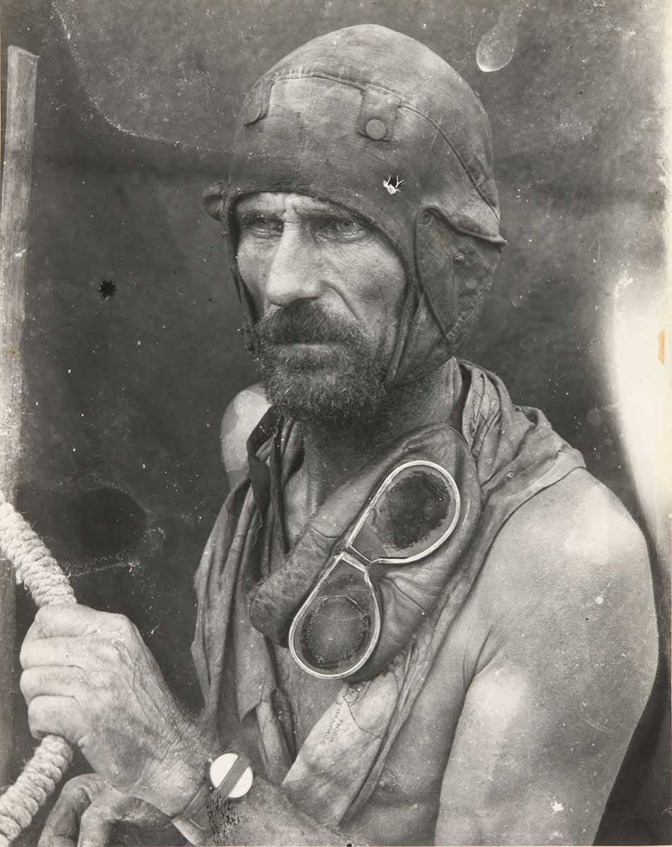 Black and white image showing a bearded and bare-chested man gazing to the left of frame. He wears a leather flying-style cap and has goggles draped around his neck. His right arm clutches a rope. - click to view larger image