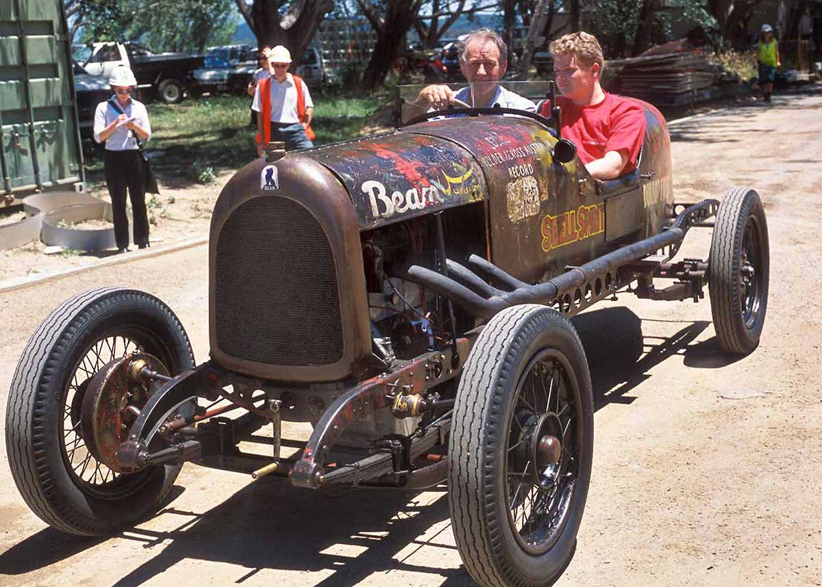 Two men sit in an open-topped car with a small glass panel windscreen. - click to view larger image