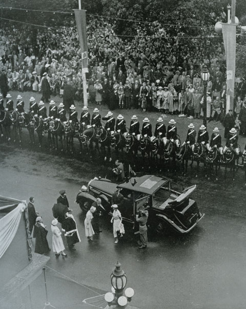 Queen Elizabeth is seen from above, walking from a vehicle toward a building. Behind the vehicle are what appear to be many police on horses lining the edge of the road upon which the vehicle is parked. A group of people wait near the building, apparently ready to greet the Queen. - click to view larger image