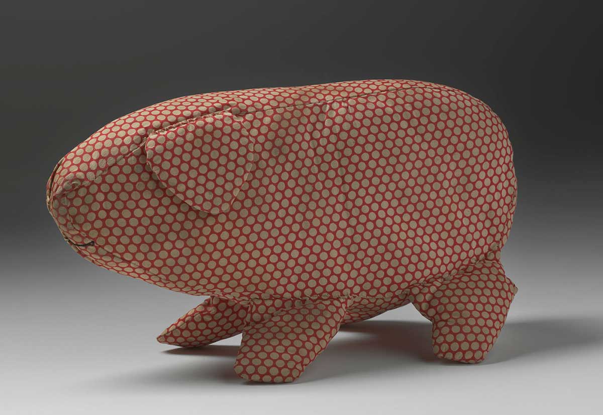 A soft toy in the shape of a pig made from off-white spotted red cotton fabric. The pig has a triangular ear and is missing its tail. A curved mouth is sewn onto the fabric in black thread.