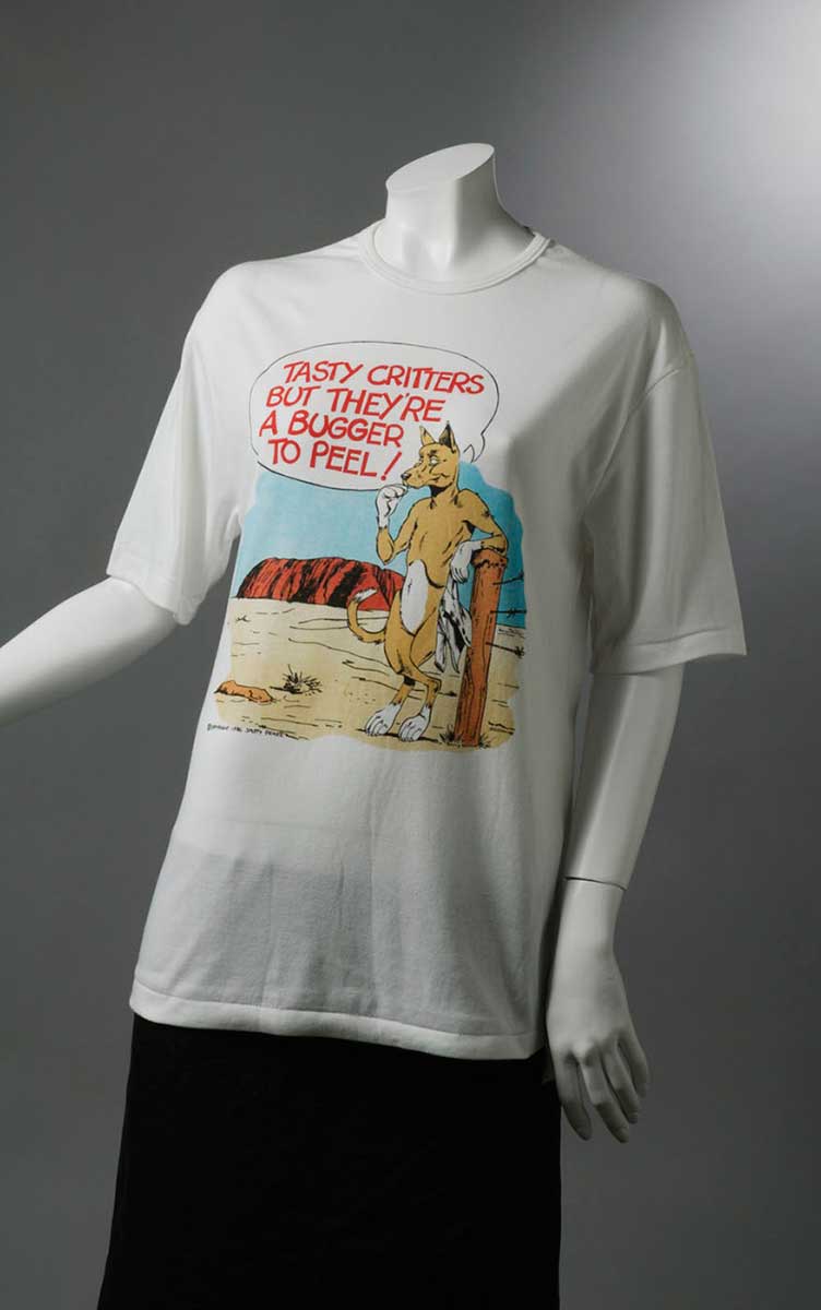 T-shirt with caricature of a dingo standing near Uluru, holding a baby's jumpsuit and saying 'Tasty critters but they're a bugger to peel!' - click to view larger image