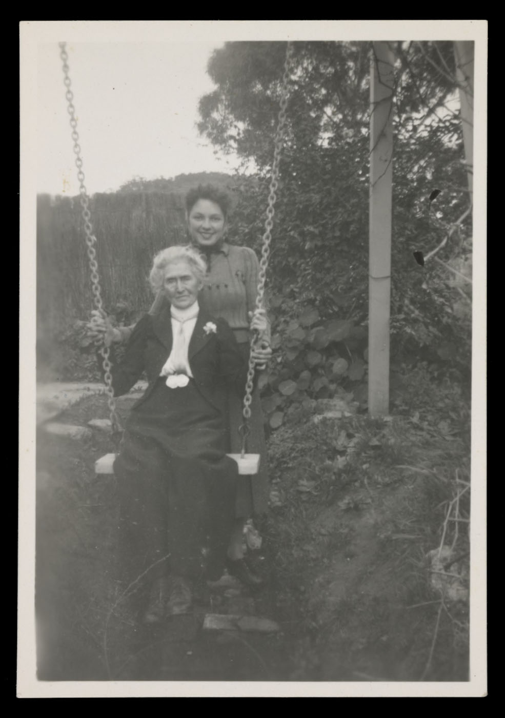 Black and white photograph showing an elderly woman sitting on a swing. A younger woman stands behind her, posing for the photograph. - click to view larger image