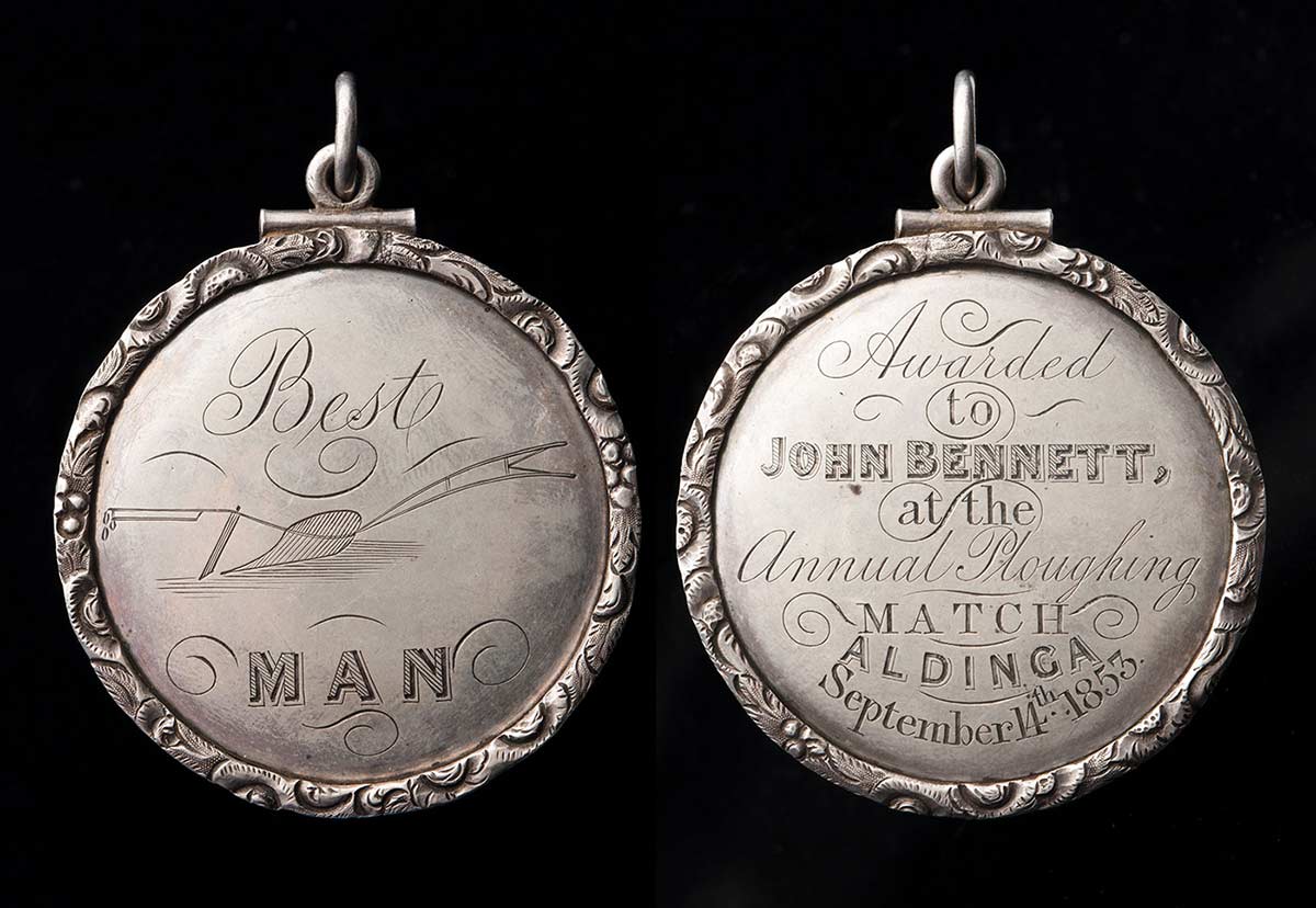 Image showing two faces of a circular medallion with a ring at the top. The face on the left is inscribed 'Best MAN' with an image of a plough. The right is inscribed with 'Awarded/to/John Bennett/at the/Annual Ploughing/Match/Aldinga/September 14th 1853'.