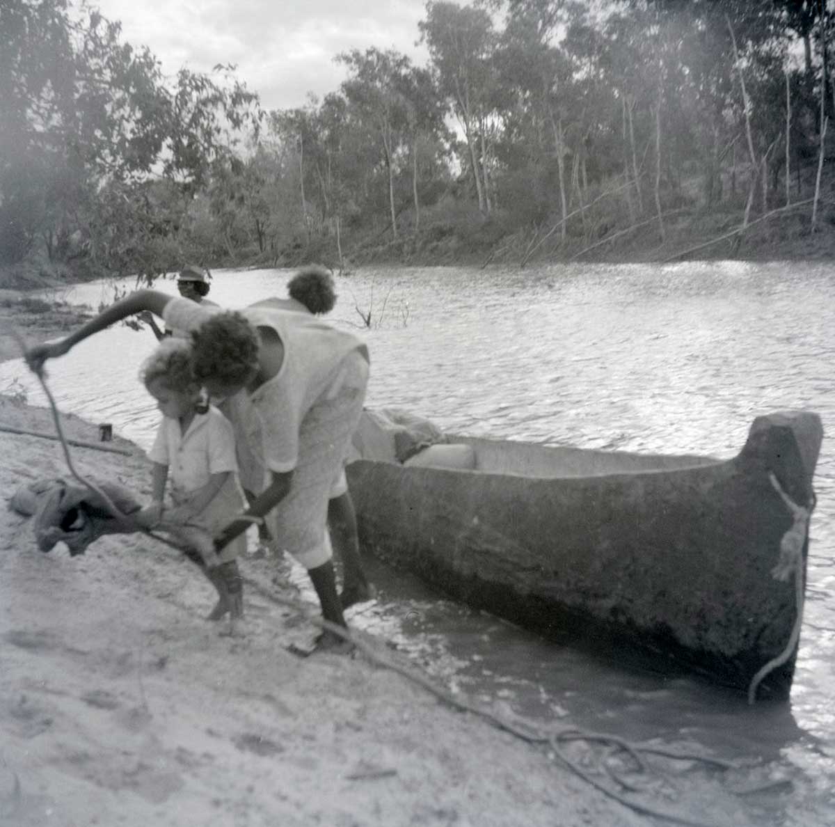 A black and white photographic negative that depicts an Aboriginal child and three adults standing on the edge of a riverbank next to a canoe. - click to view larger image