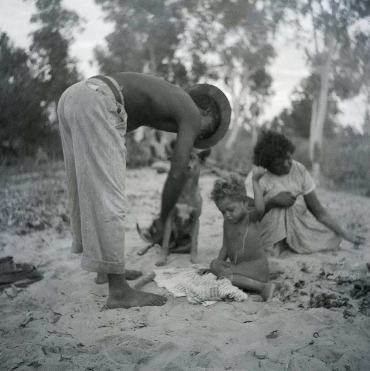 A black and white photographic negative that depicts an Aboriginal man wearing a hat bending over next to a child with a woman and dog sitting in the background. - click to view larger image
