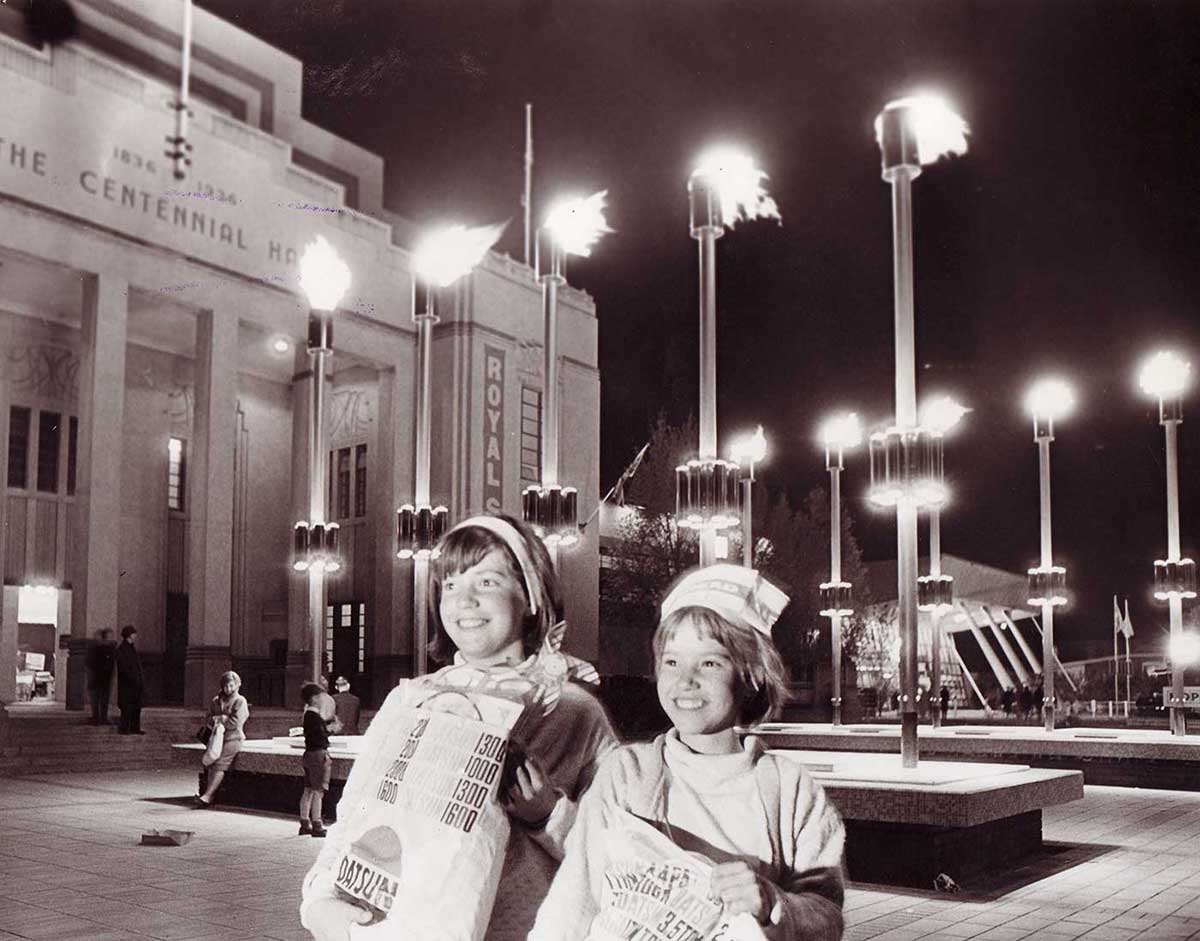 Black and white photo showing two girls posing with show bags outside the Centennial Hall at night. - click to view larger image
