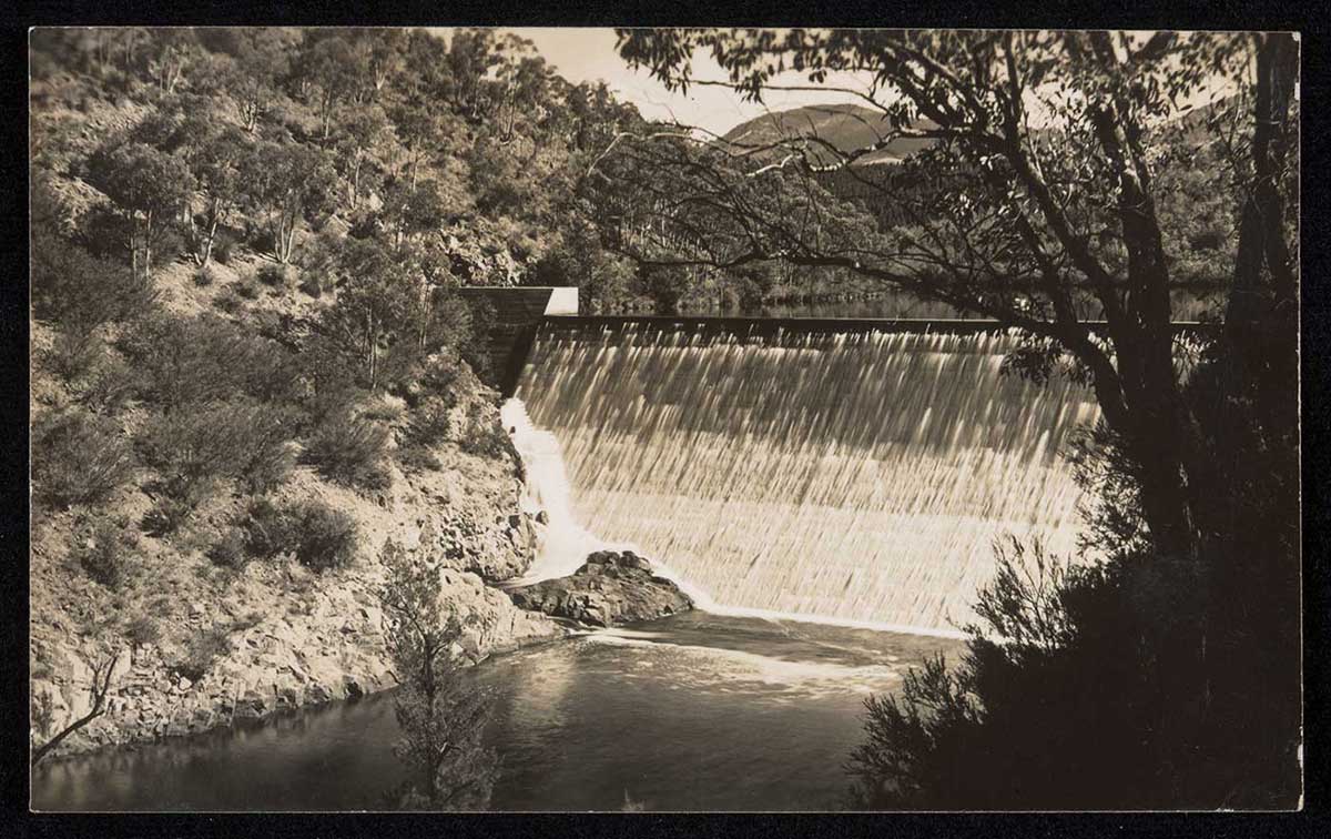 Black and white postcard showing a photograph of a concrete spillway, above a waterway. Gum trees line the steep bank on the left side. - click to view larger image