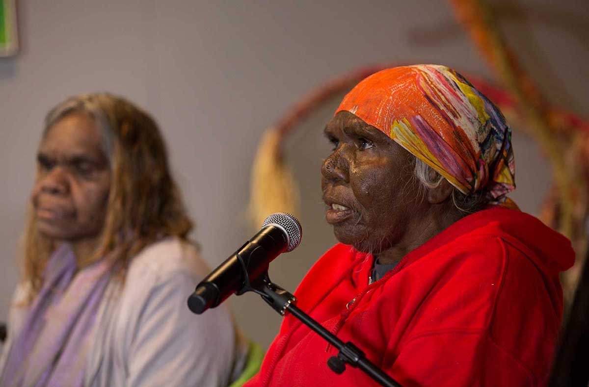 A woman speaks into a microphone, while another woman watches on. - click to view larger image