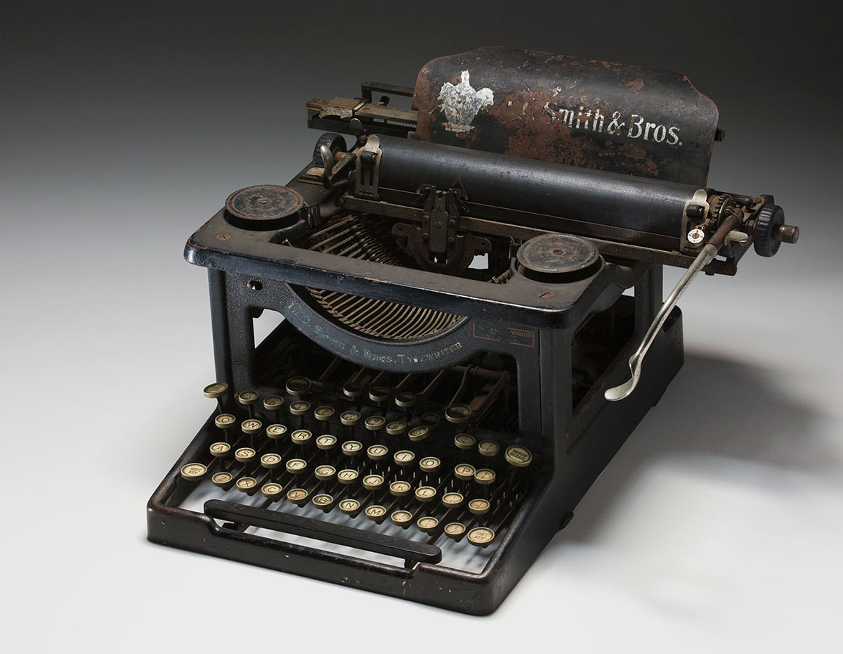 Black painted, steel framed, 'visible' typewriter with circular white pre-decimal keys. 'L.C. SMITH & BROS TYPEWRITER' is printed along an inverted arch across the front, which cradles the typeface heads. It is rusty and the ribbon is missing. - click to view larger image