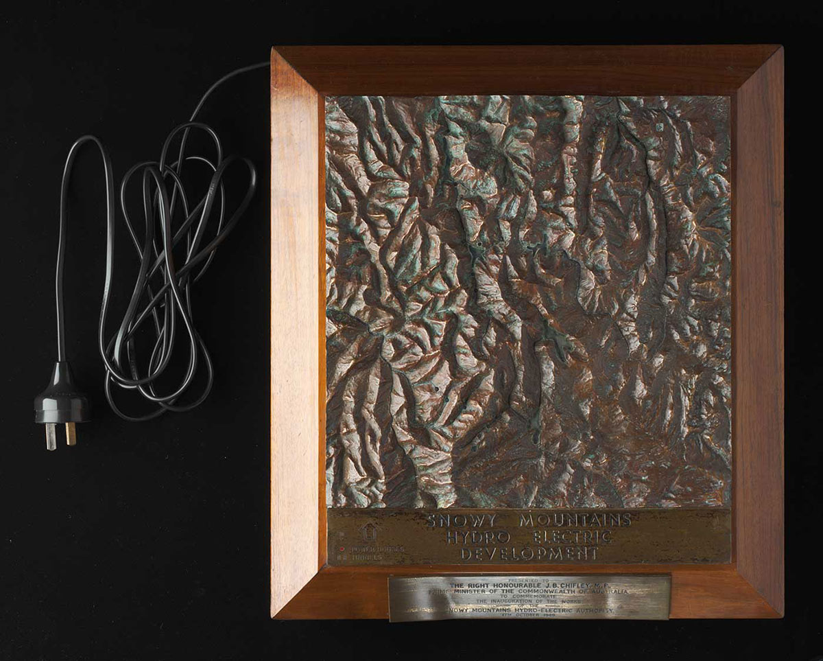 A bronze topographic plaque depicting the topography of the Snowy Mountains, mounted on a brown wooden plinth with the engraving ‘SNOWY MOUNTAINS / HYDRO ELECTRIC / DEVELOPMENT’ and a black electrical cord coming from the top left of the plaque. - click to view larger image