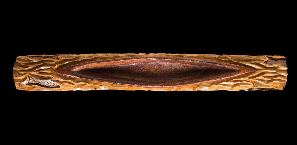 A lacquered wooden pole with an elliptical shaped carving across the top depicting a canoe which is surrounded with carved fish and wave-lines. - click to view larger image
