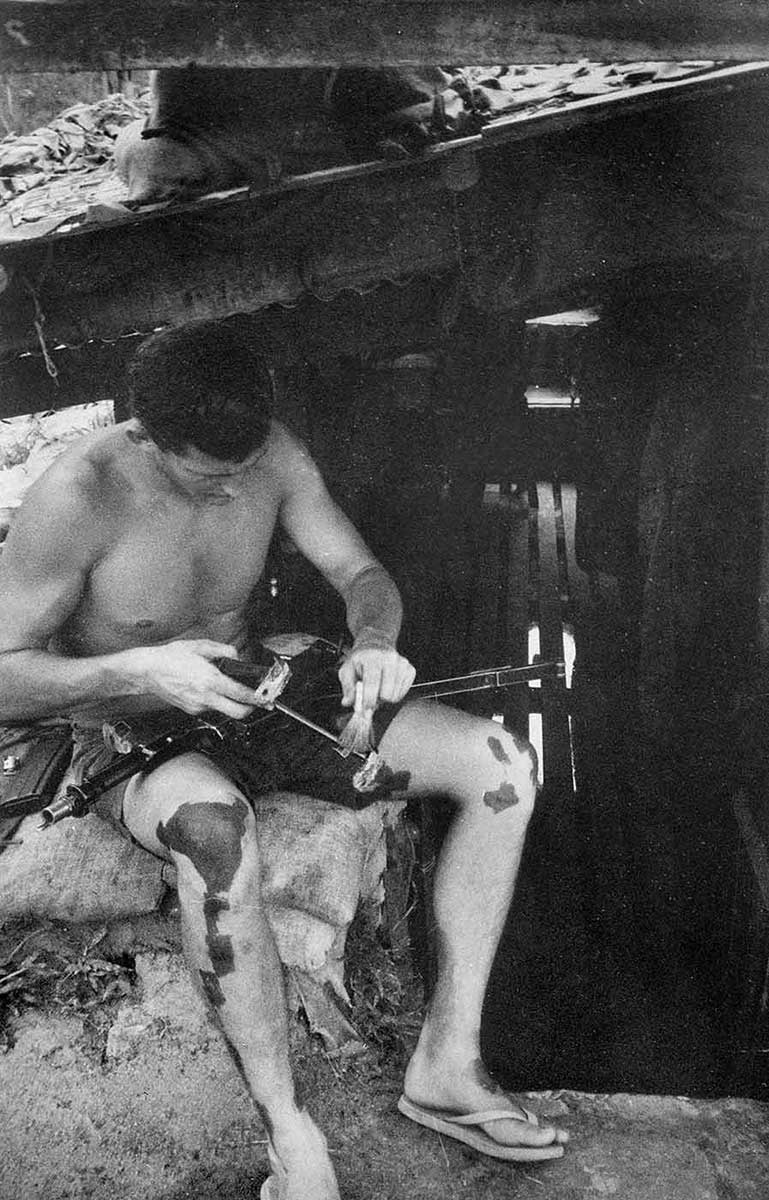 Young man wearing just shorts and thongs sitting on sandbag, cleaning gun. His knees are covered in dark patches. - click to view larger image