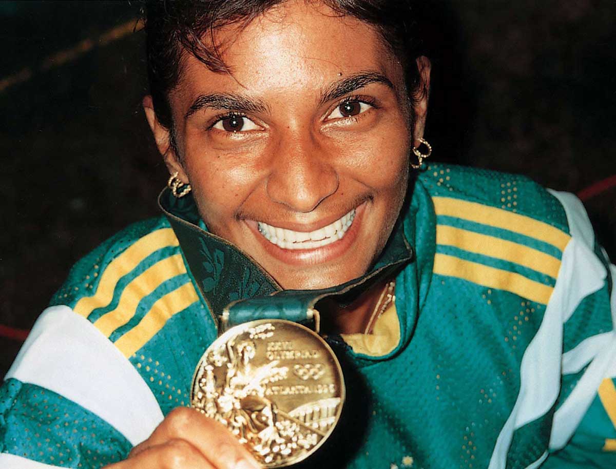 A smiling woman wearing green and gold tracksuits holds a gold medal.