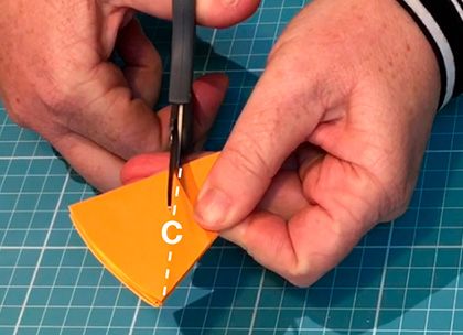 A piece of orange paper which has been partially folder over. Fingers from one hand hold down one side of the paper while the other hand holds a pencil which points to a corner with the capital letter A in white font.
