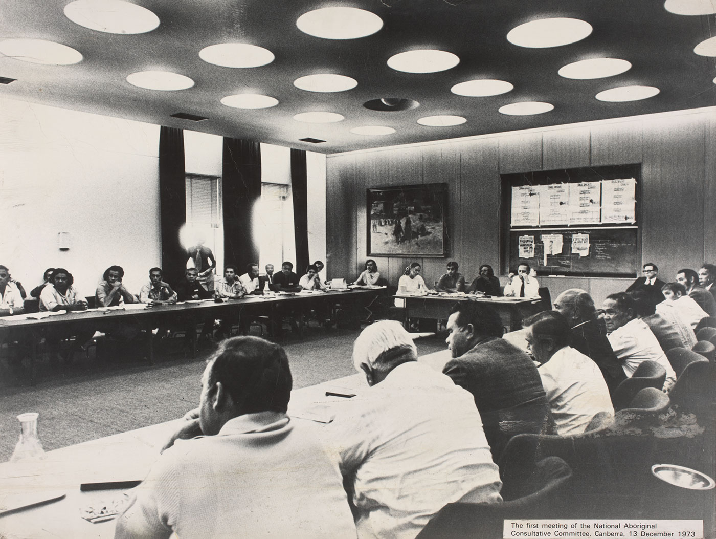 Black and white photo showing a meeting room with numerous Indigenous people sitting at tables arranged in a rectangular formation.