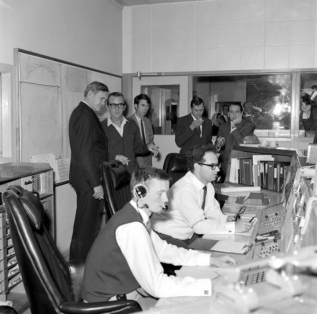 A black and white photo taken in 1969 featuring the then Prime Minister Gorton accompanied by several other men. They are watching on while two men, who are wearing headsets and seated, appear to be at a workstation. - click to view larger image