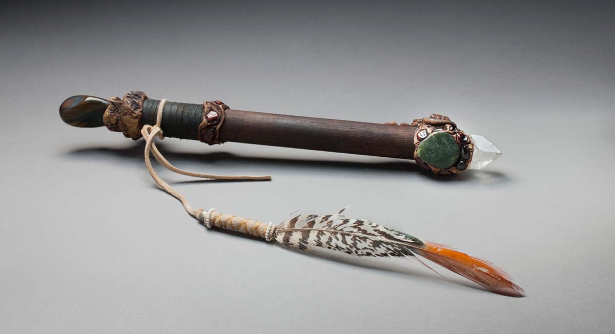 A cylindrical piece of wood with decorative bands into which are set polished semi-precious stones. The stick has a polished agate pebble at one end and a pointed piece of rock crystal at the other. The stick has a leather string attached with a silver bead followed by a decorative section with white beads and ends with a long feather. - click to view larger image