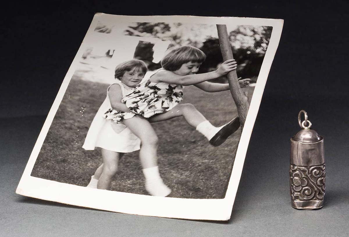 A black and white photograph of two girls climbing a tree. The girls are both wearing summer dresses, and there is a washing line in the background. Second object is a silver coloured metal charm, which is cylindrical in shape, and features a floral pattern. The charm is slightly misshapen. - click to view larger image