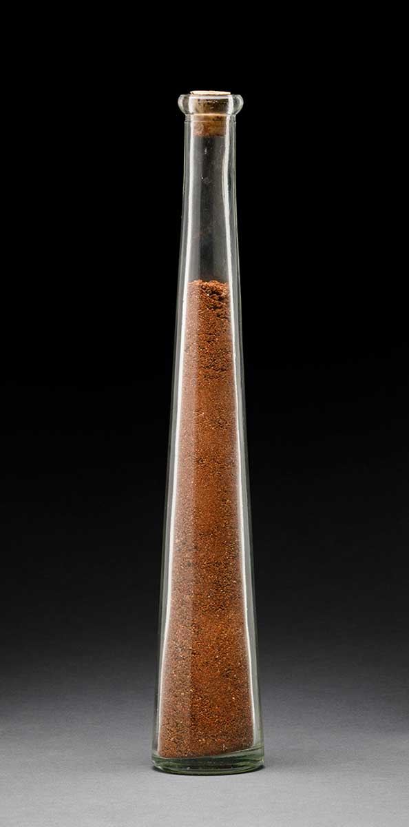 A glass vial with a cork in the top and the number '10' imprinted on the base. Inside is red-brown coloured soil and rocks. - click to view larger image