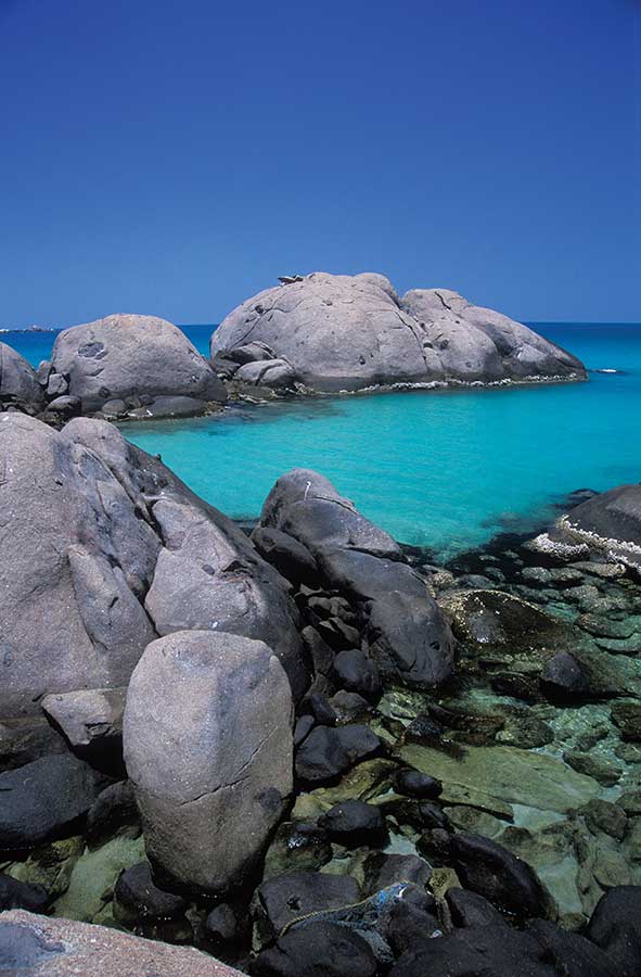 A colour photograph of a section of coastline. In the foreground are various rocks. Some are dark grey and flat, while others a lighter grey and more rounded. In the middle ground is a small inlet of water. Beyond it are two light grey large boulders. Parts of the horizon can be seen beyond the boulders. The blue cloudless sky is in the upper third of the photograph. - click to view larger image