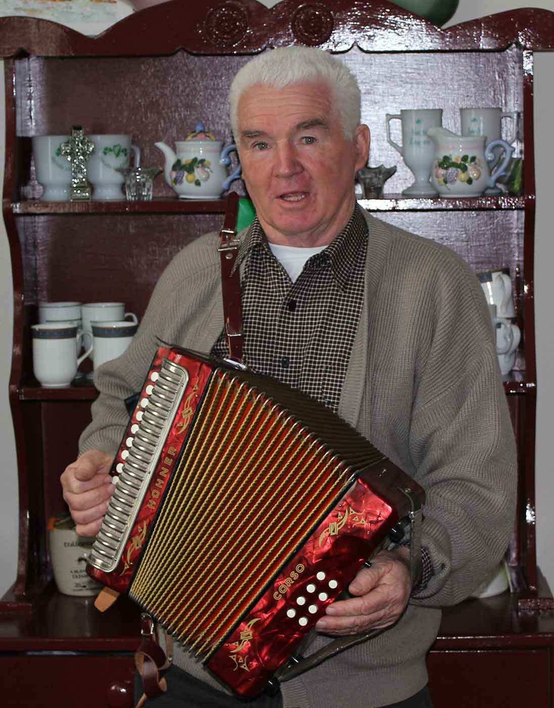 An elderly man is playing an accordion in front of a kitchen dresser. - click to view larger image