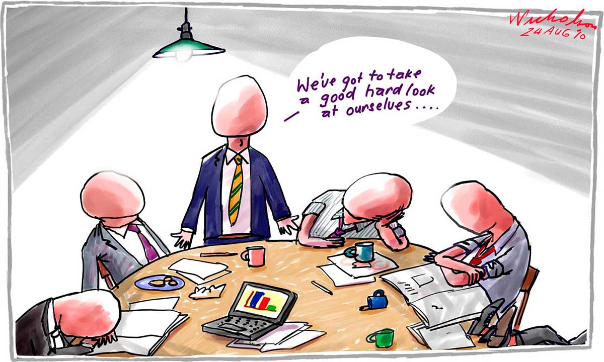 A colour cartoon depicting six men around a table. Four are sitting; one rests his head on the table. One has his feet on the table; that is all that can be seen of him. All of the entirely visible men have featureless heads, with no faces, ears or hair. On the table are papers, a laptop computer, pens and some coffee mugs. One of the men stands and says 'We've got to take a good hard look at ourselves ...' Above him, a single lightbulb under a shade shines down upon the group. - click to view larger image