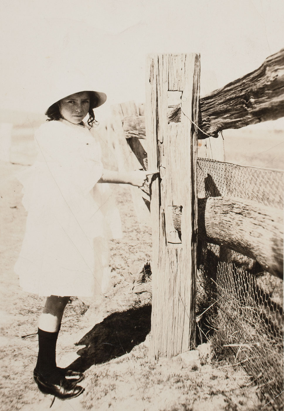 Black and white photo of a young girl standing adjacent to and pointing at an old wooden fence. - click to view larger image