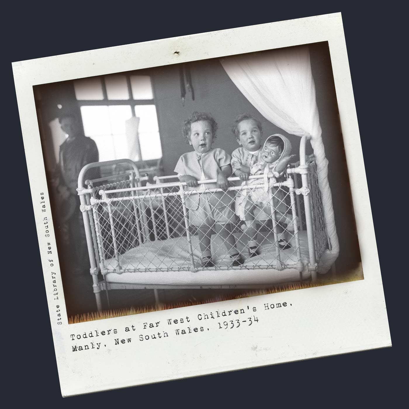 A black and white Polaroid photograph of two small children in a metal cot. The children are standing facing the camera and holding the raised side rail. One of them is holding a large doll. There is a man standing in the background who is looking towards the children. Typewritten text underneath reads 'Toddlers at Far West Children's Home. Manly. New South Wales. 1933-34'. Smaller text, on the left-hand side of the image, in a vertical direction, reads 'State Library of New South Wales'. - click to view larger image