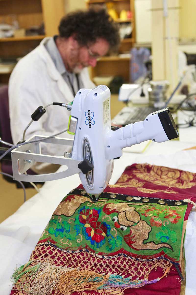 A conservator using XRF to identify the metallic make-up of threads and fabric in a Chinese processional costume from the 1880s. - click to view larger image