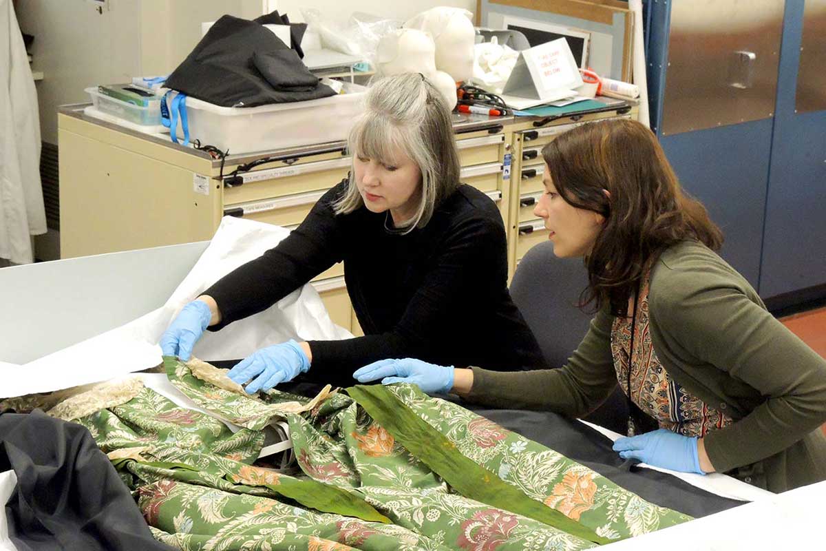 Two female conservators work on a dress with intricate pattern and lace detail in a laboratory.