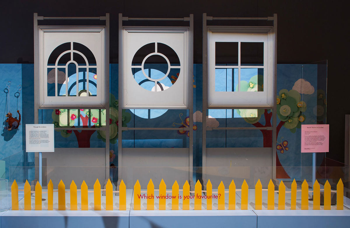 Exhibition display featuring three window frames. One is in the shape of an arch, one is in the shape of a circle and one is in the shape of a square. In the foreground is a yellow picket fence and exhibition text on Plexiglas which reads ‘which window is your favourite?’ - click to view larger image