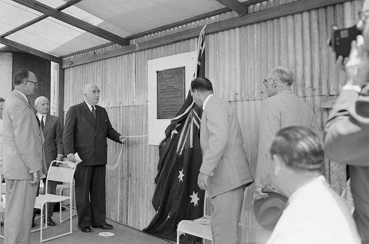 Black and white photo of Prime Minister Robert Menzies unveiling a plaque covered by the Australian flag and mounted on a wall that is constructed from corrugated tin roof panels. A group of men in business suits watch the unveiling.