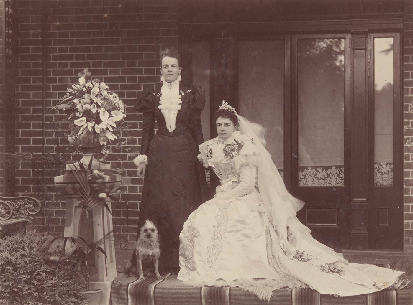Black and white photo of a woman in a wedding dress and veil, seated. A woman in a dark dress stands at her left. A small terrier dog sits at her feet. - click to view larger image