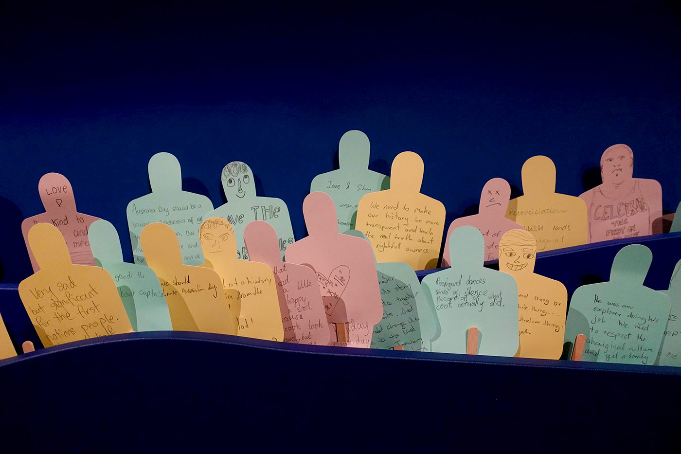 Exhibition display featuring multiple pieces of coloured paper cut out in the shape of people - some with faces - with hand written messages.