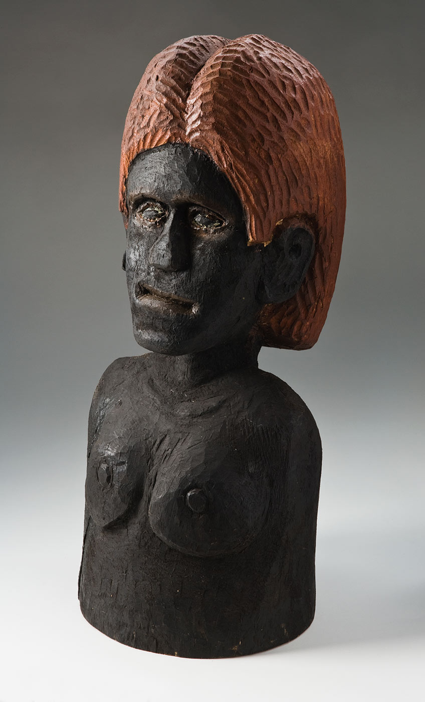A carving of a nude female torso made from a solid piece of cylindrical wood and pigmented. Most of the carving is coloured black with the facial and chest features carved lightly in relief. The eyes have an outline of cream paint and the nose is flattened. The shoulder length hair is coloured orange brown, and is covered with short vertical grooves. - click to view larger image