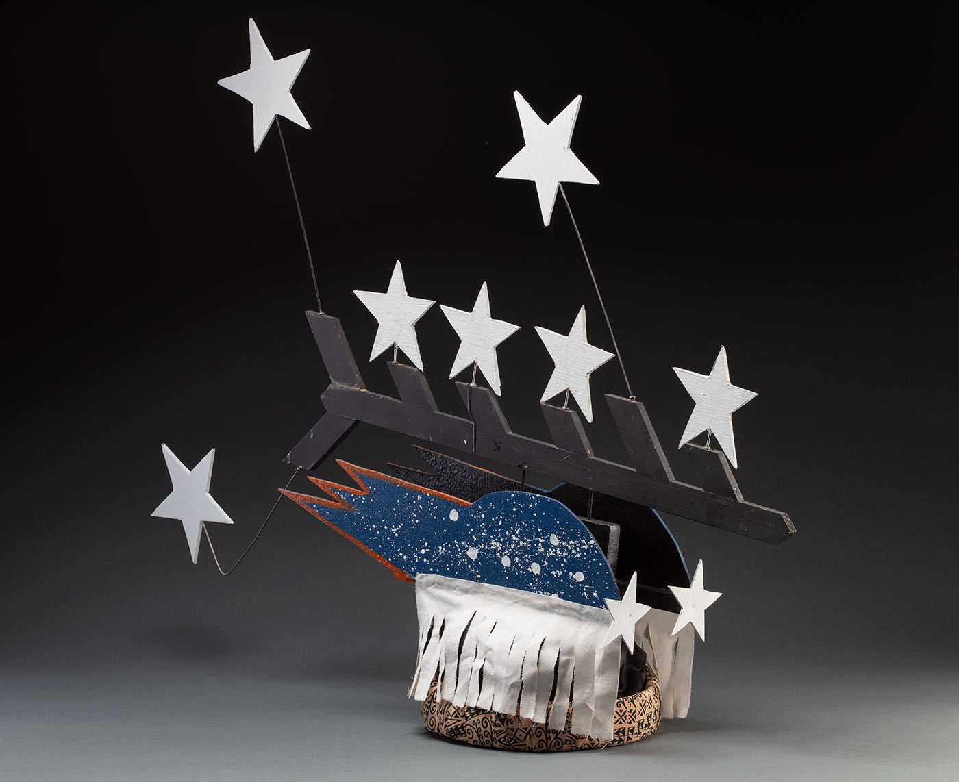 A headdress featuring seven stars protruding from a black painted piece of board forming half a fishbone pattern. It is connected to two pieces of board painted blue with white splatter and seven stars on each side with a white fabric fringe at the bottom. It is all attached to a fabric wrapped headband in the centre.
