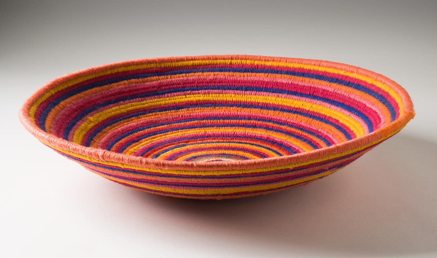 A multi-coloured circular coiled bowl-shaped basket made of yarn and plant fibre. The centre of the basket is in turquoise yarn followed by horizontal stripes of yarn in orange, blue, yellow, dark pink, red, green, brown, and pale pink. - click to view larger image