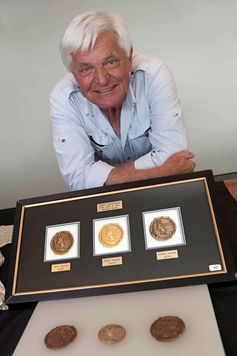 Colour photograph of a man resting with folded arms on a table. In front is a framed set of three circular medals. Another three medals are in the foreground. - click to view larger image