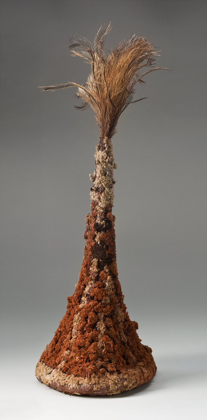 A tall conical headdress made of plant fibres attached to a firm circular ring at the base and covered with pigmented fluffy plant fibre with feathers at the top. The ring at the base is pigmented red and the plant fibre stalks are attached around it. The fibres are bunched together towards the top and wrapped with maroon coloured string. The plant stalks are covered with fluffy plant fibre which is decorated with red pigment in vertical stripes on the bottom half and cream at the top. At the top of the headdress is a bunch of fluffy and spiky brown, tan and grey feathers. - click to view larger image