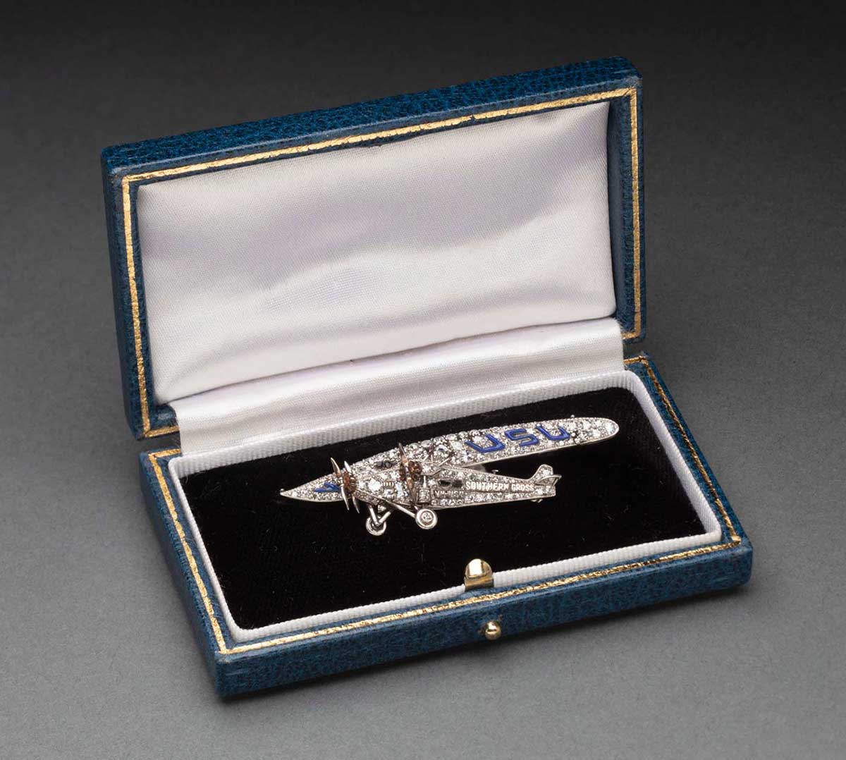 A diamond and platinum brooch in the form of an aeroplane in a blue rectangular box with gold detailing and a push button latch. On the brooch there are sapphires forming the windows and blue enamel on the wings. The side of the plane reads 'VH-USU / Southern Cross'. The interior of the box lid is covered in white coloured fabric and the base is covered in black coloured fabric. - click to view larger image