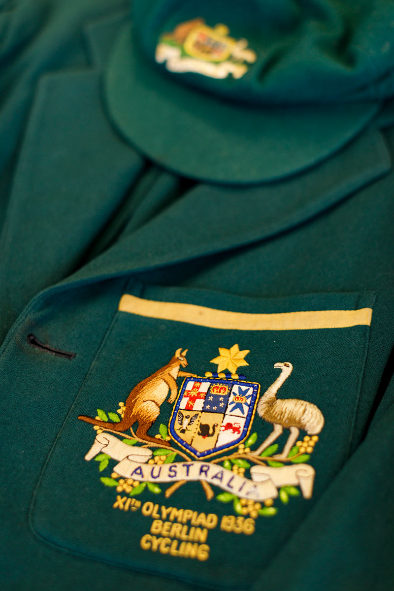 Close up of a green uniform with yellow detailing. On the breast pocket is an embroidered crest of a kangaroo and emu on either side of a shield, and the text: IX-OLYMPIAD / AMSTERDAM-1928. - click to view larger image