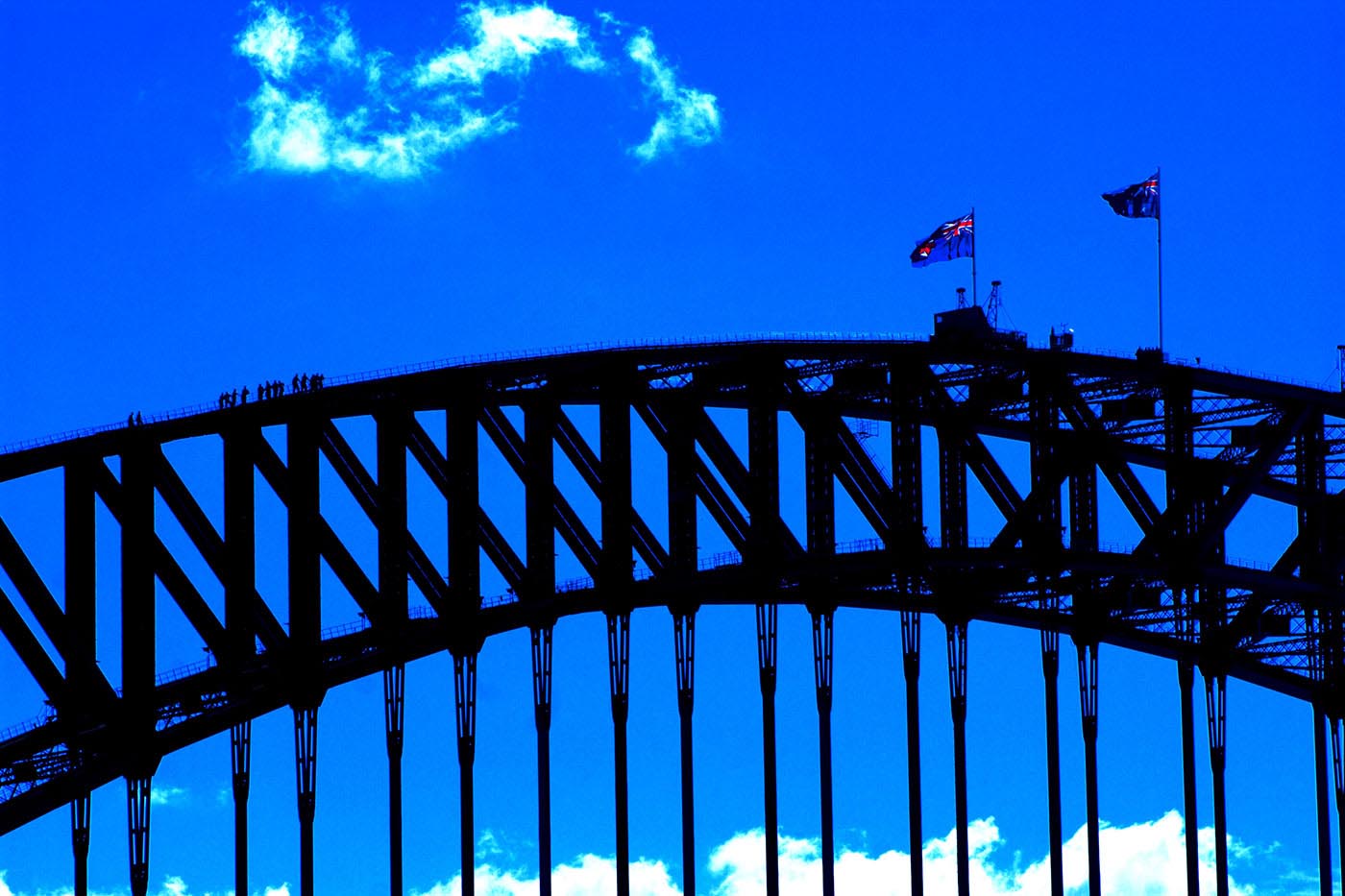 Colour photograph showing the top of the Sydney Harbour Bridge. Two flags fly from the top centre of the arch. A group of climbers, dwarfed by the structure, approach the top of the arch from the left. - click to view larger image