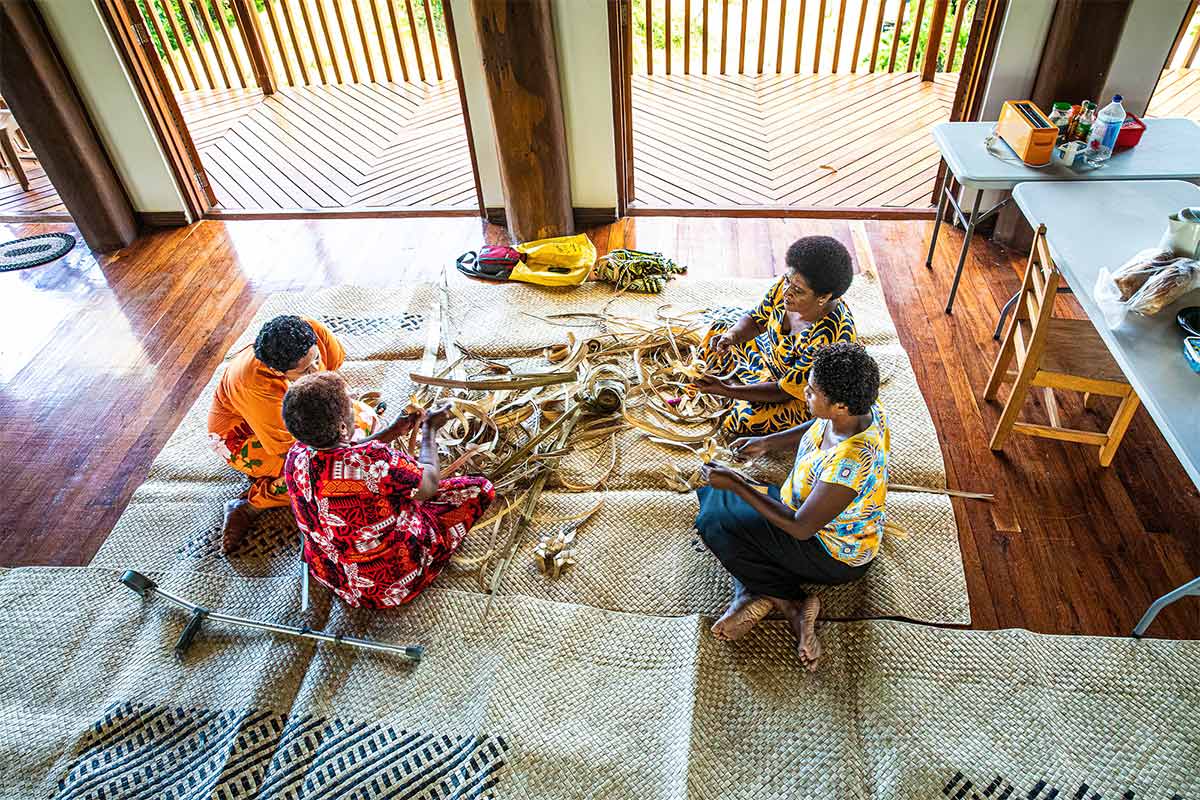Colour photograph featuring a group of four women seated on the ground on large woven mats, making artworks with natural materials. - click to view larger image