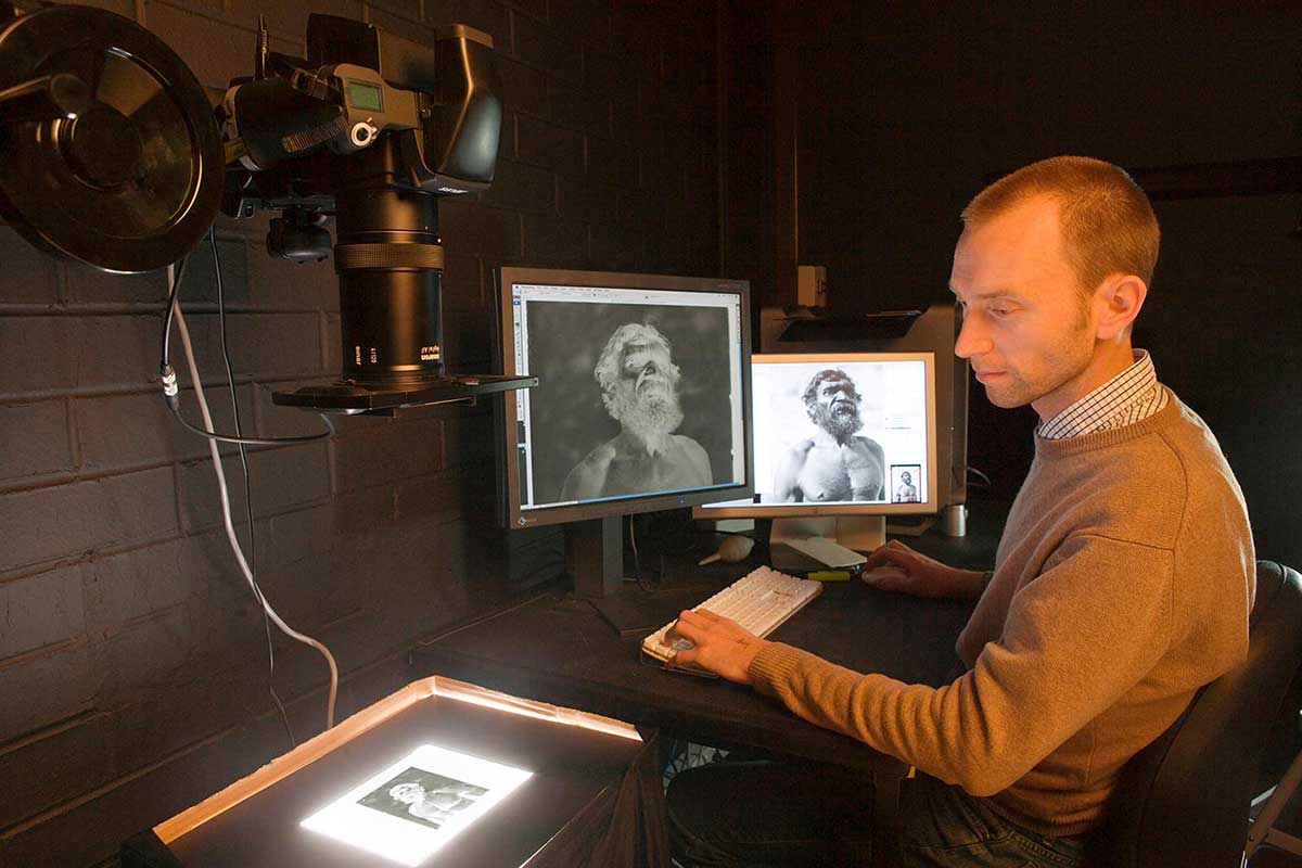 One of the Museum's photographers digitising the Basedow collection. The photographer sits at the right of the image facing to the left. He is in a darkened corner of the Museum's photography facility. The table at which he sits has a keyboard, computer mouse and two computer flat screens on it. To the left of the image is a digital camera mounted vertically above a flat surface. The camera is projecting a negative image of an Aboriginal man onto the surface. Digital negative and positive images of the man can be seen on the computer screens in front of the photographer, who looks at the negative image on the flat surface. The photographer appears to be making adjustments to the digital images via keyboard and mouse commands. - click to view larger image