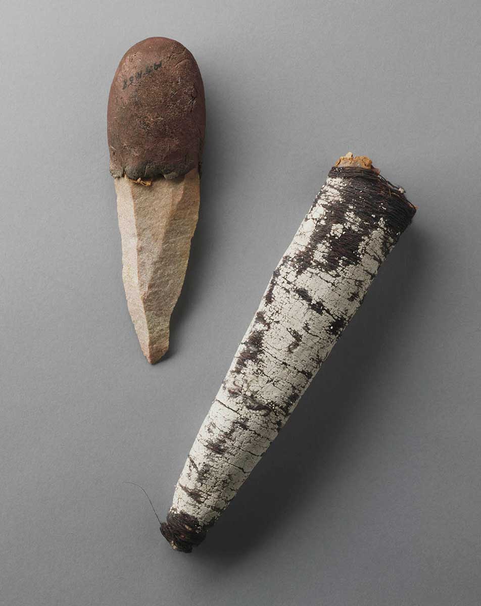 A stone flake-bladed knife with a pigmented melaleuca bark knife sheath. The knife blade has three faces on its top surface and a rounded natural resin handle. The melaleuca bark sheath has been covered in string made from natural fibres. Its surface has been painted with white ochre, which is cracked and missing in places. - click to view larger image