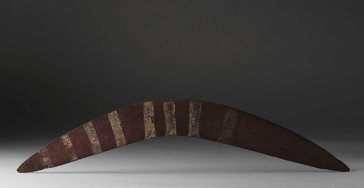 A boomerang, presenting its curved top surface, photographed standing upon its ends. The ends taper to points. The top surface has eight white bands on it, running across the width of the surface. The bands are faded and grouped toward the middle and right-hand side of the boomerang. - click to view larger image