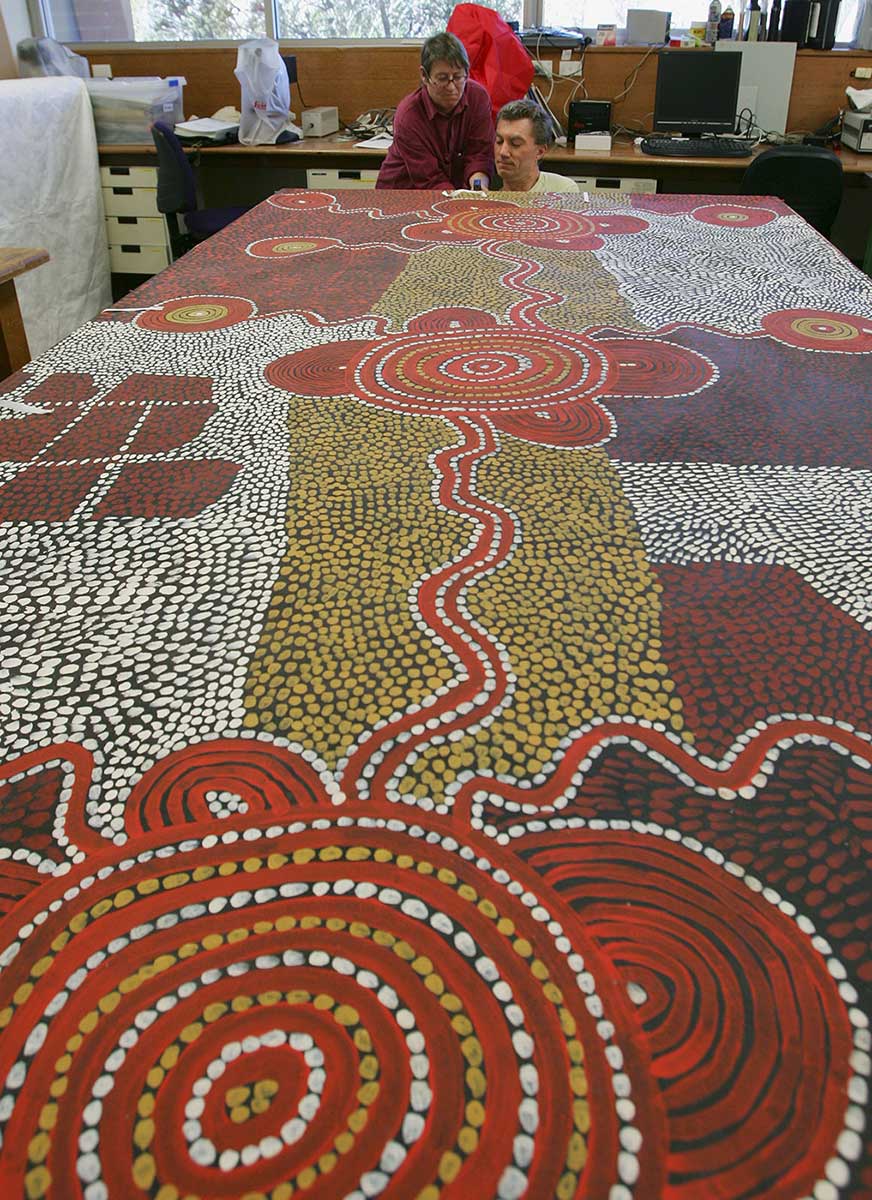 Two conservators inspect far end of large Yumari 1976 dot painting by Uta Uta Tjangala. - click to view larger image