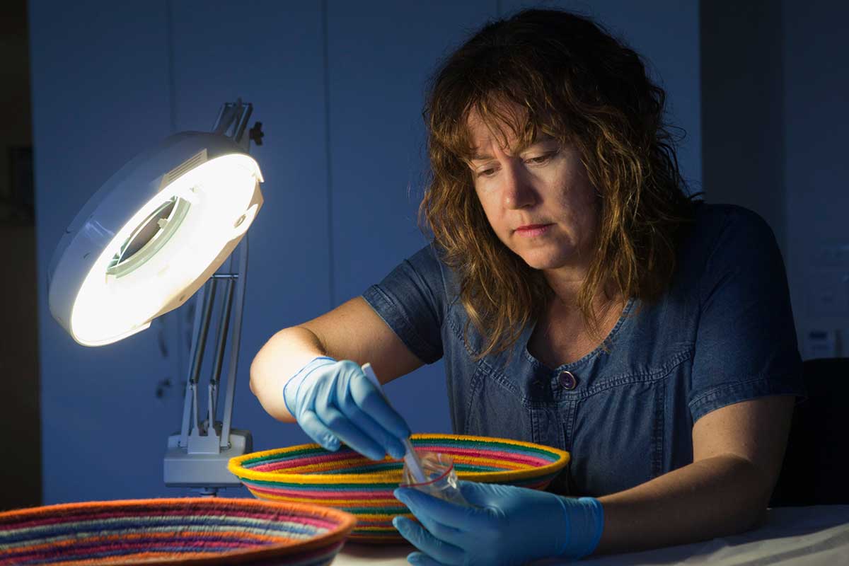 A colour photograph of a woman sitting at a table with two brightly coloured baskets on top, lit by a bright lamp. The woman, who wears blue gloves, uses tweezers to place small objects in a plastic bag.