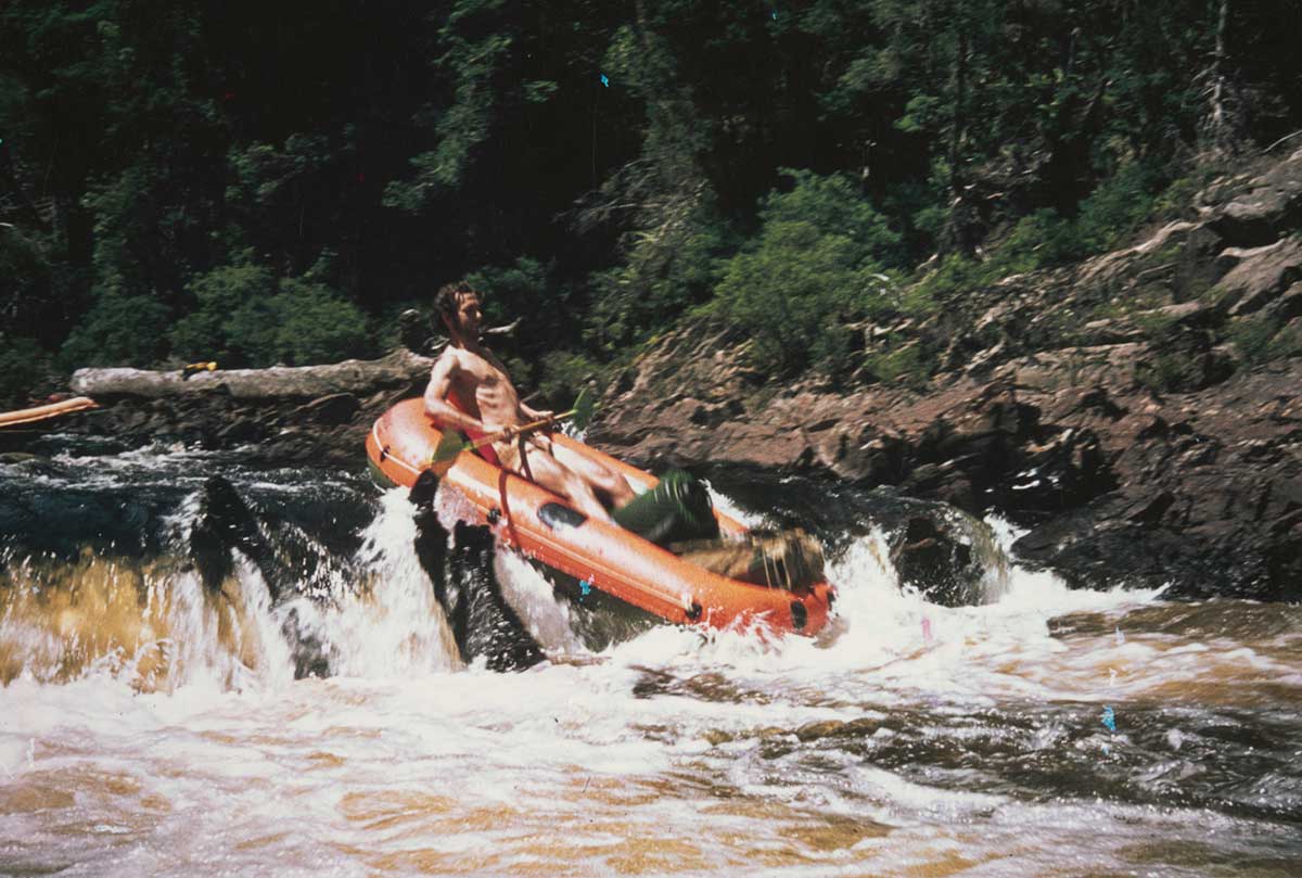 A colour photograph of Bob Brown going down a rapid on a river in an orange inflatable raft. He has a green and a brown bag in the front of the raft and is holding the paddle. He has a beard and is wearing shorts.
